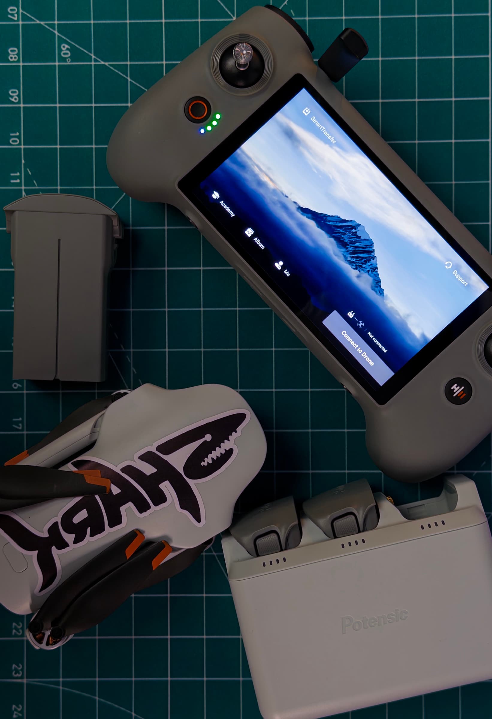 Top-down flat lay of the folded ATOM 3, its touchscreen controller, a spare battery and the Potensic charging hub on a cutting mat