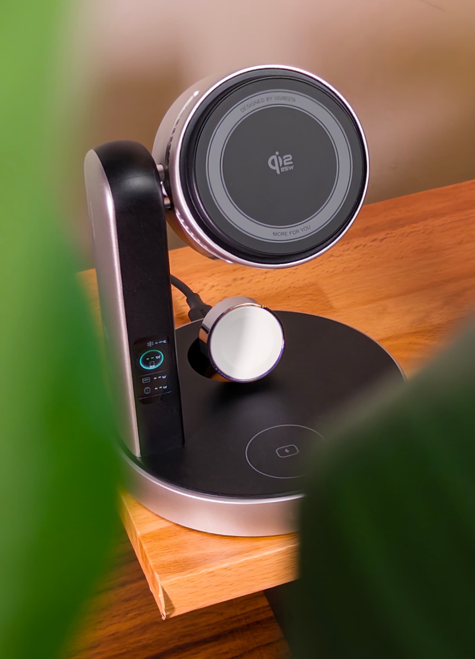 MagFlow Pro on a wooden desk seen through blurred plant leaves, with its Qi2 pad, watch charger and status display